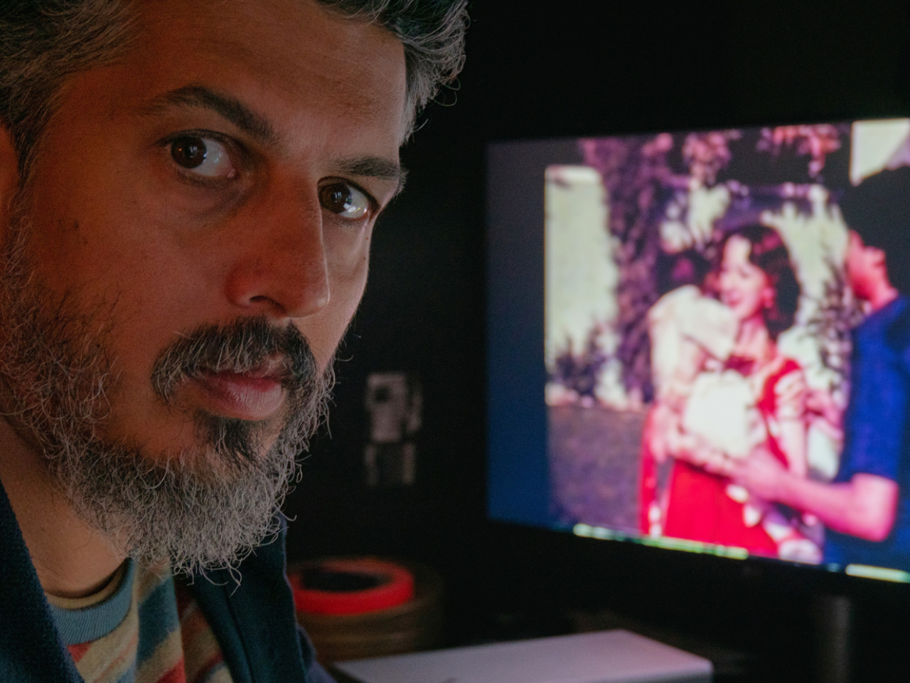 Director Sifian Khan turns to face the camera. Just over his shoulder, a TV screen shows a couple wearing bright colors.