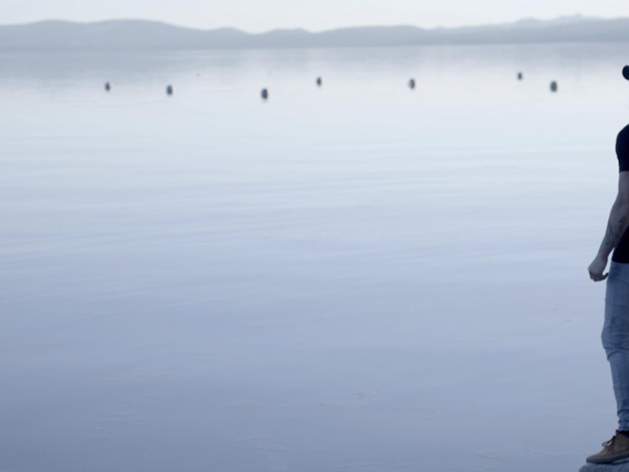 A production still from the film Momentum. A person stands to the extreme right of the frame and looks out at a lake.