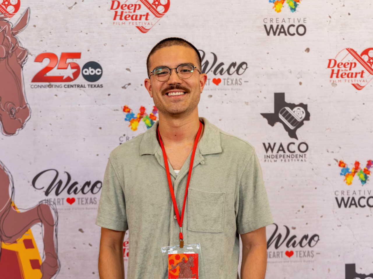 New Day Filmmaker Jake Solomon faces the camera and smiles. He wears a green shirt and is in front of a banner with logos.