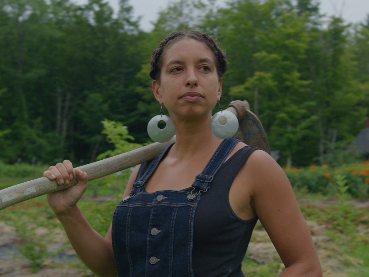 Black woman in overalls stands center with garden hoe over her right shoulder