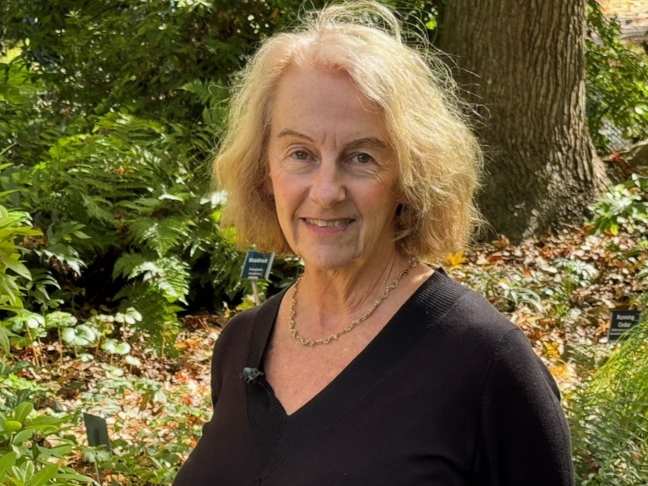 Joanne Hershfield, a woman with bright blond hair and wearing a v-neck black blouse, stands in a wooded area, smiling at the camera.