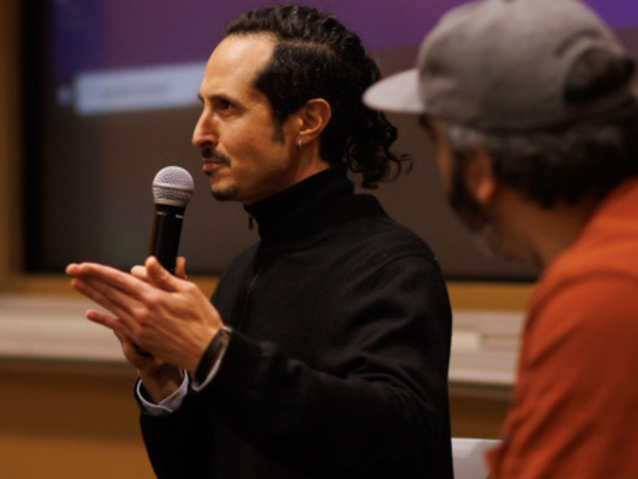 Filmmaker Joel Fendelman, a white man with long, curly black hair, stands at the front of an auditorium addressing an audience. He speaks into a microphone, and another man looks on from beside him.