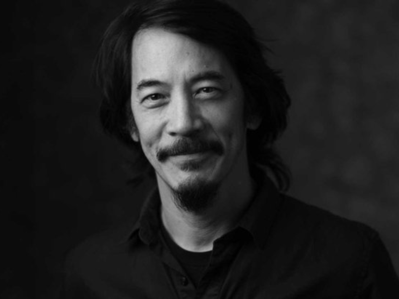 Black-and-white portrait of New Day Films Director Mark Decena. He is smiling at the camera with dark hair, a mustache, and goatee against a dark background.