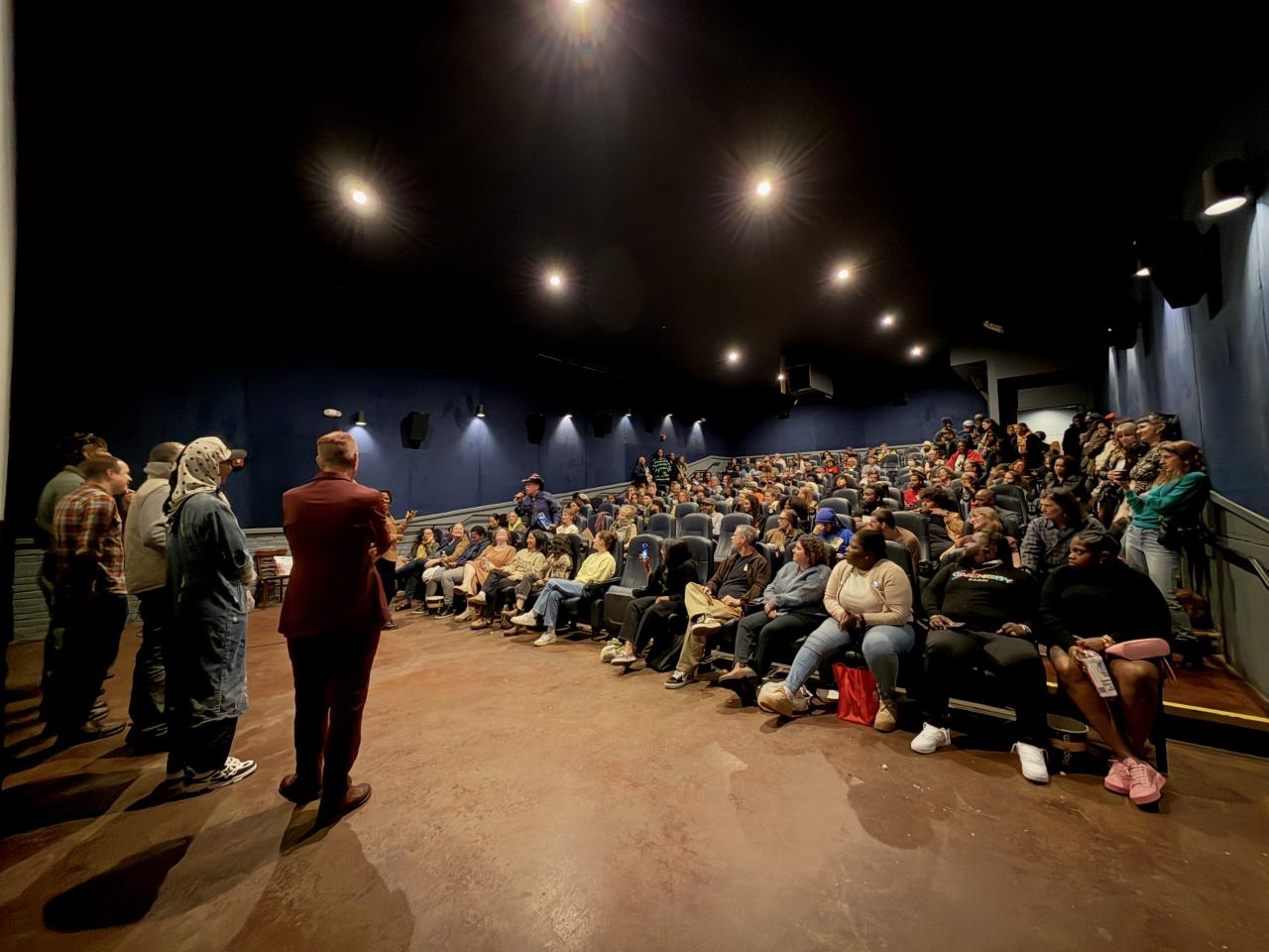 Five people stand at the front of a small theater speaking on a panel after a New Day Films screening. People of a range of ethnicities and genders fill nearly all of the seats, and more line the walls.