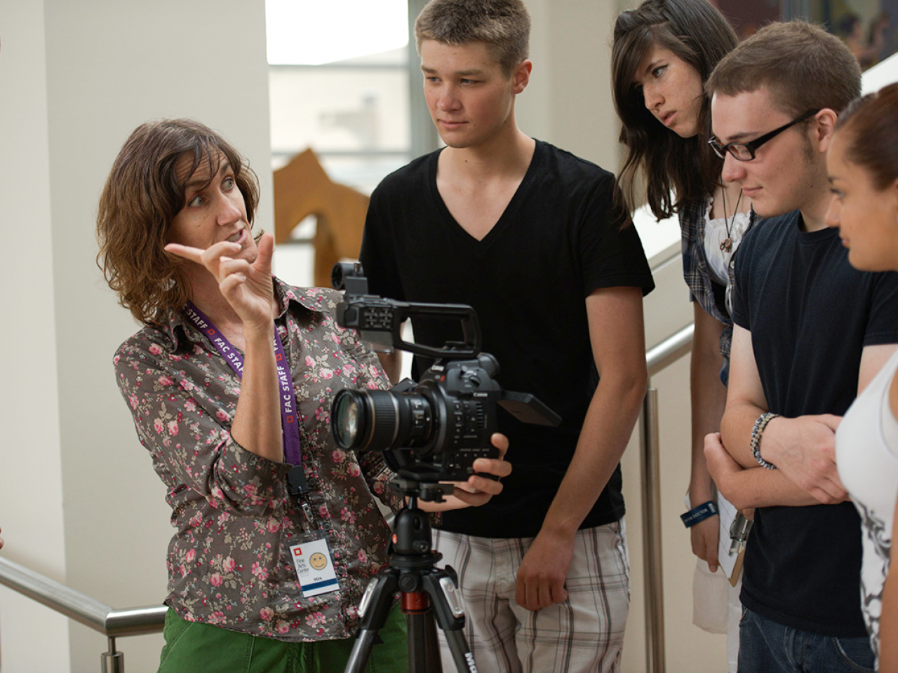  Suzan Beraza, a Latina woman, addresses a group of attentive students around a camera on a tripod. She points to their subject matter off-screen while looking at the students.