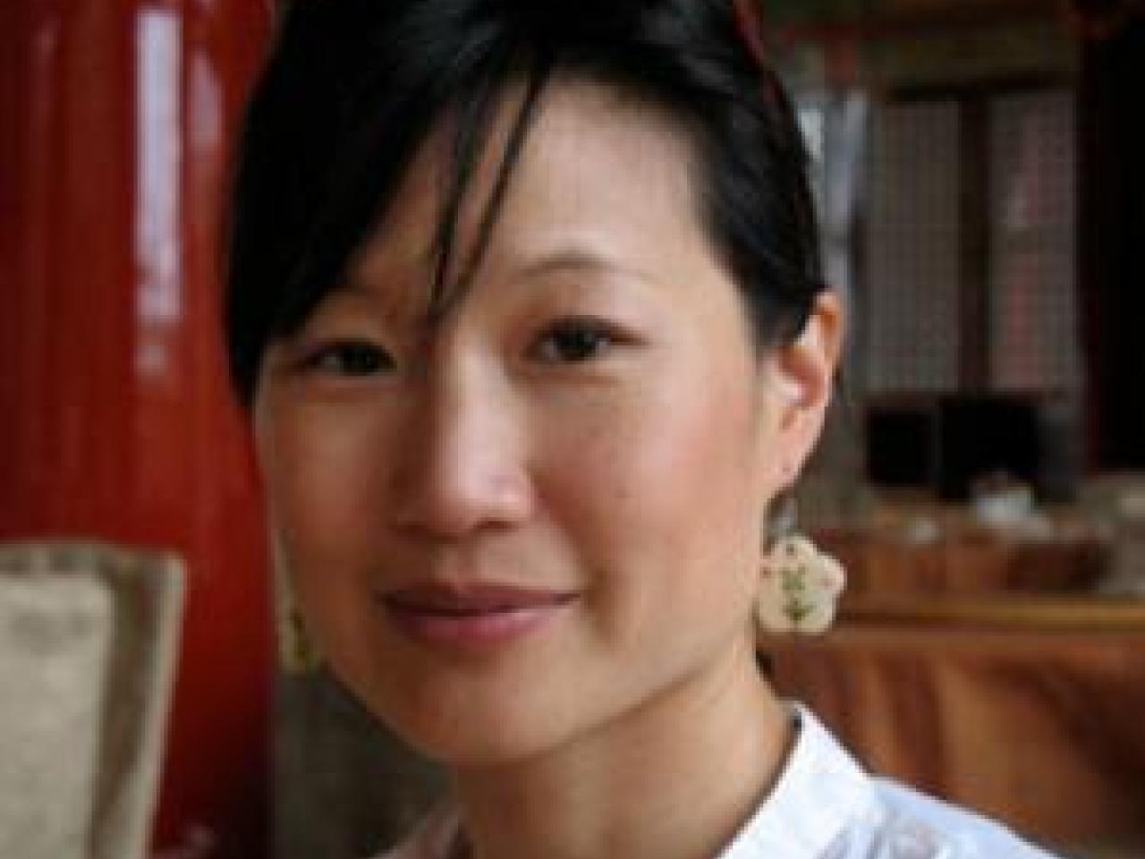 A headshot of filmmaker Debbie Lum smiling softly towards the camera. Her hair is tied back and her side bangs fall across her forehead. She is wearing a white collared shirt, red sunglasses, and flower-shaped earrings.