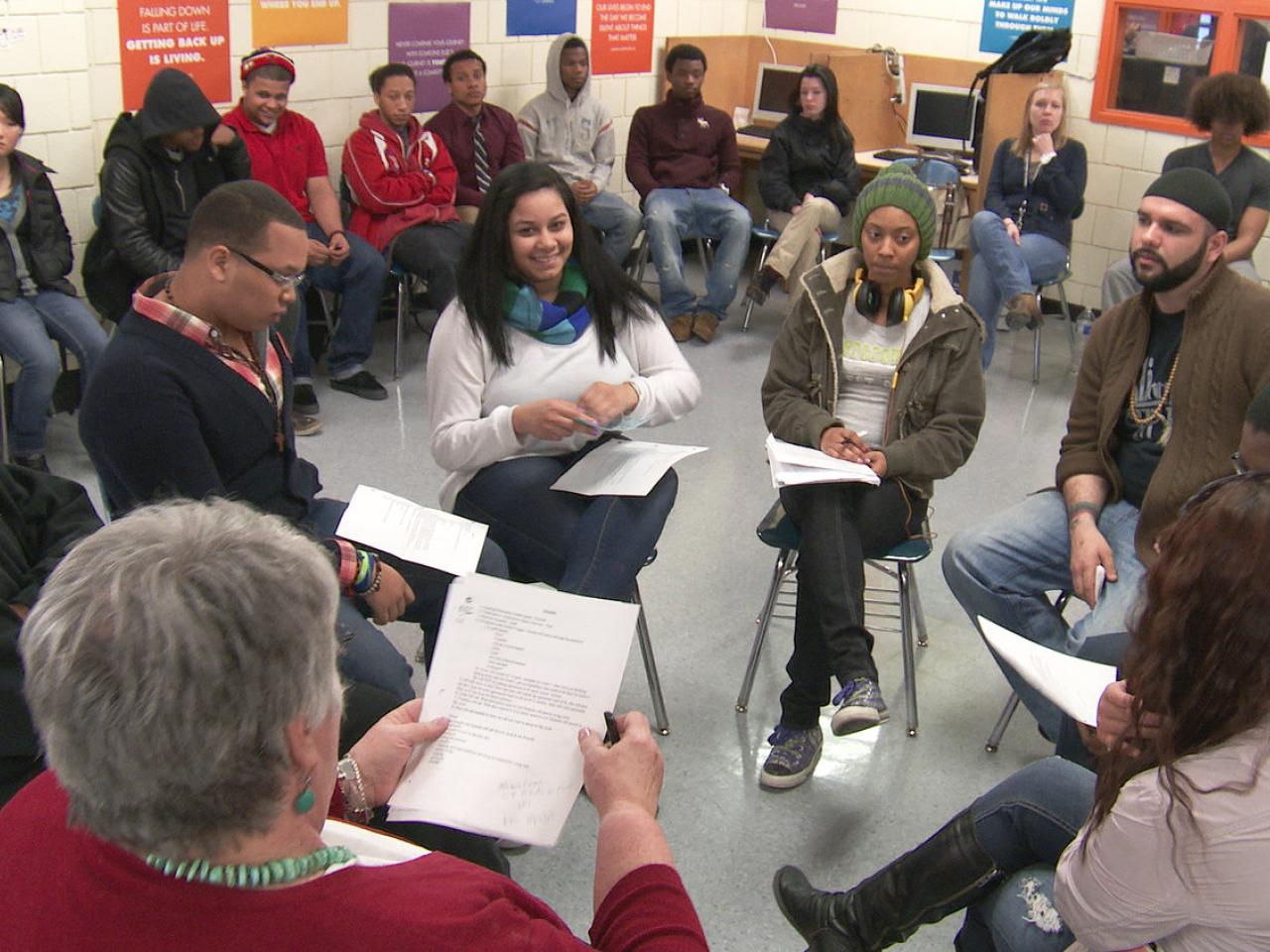 A multiracial group of people sit in a circle at a restorative justice training. More people sit in chairs lining the walls around the inner circle.