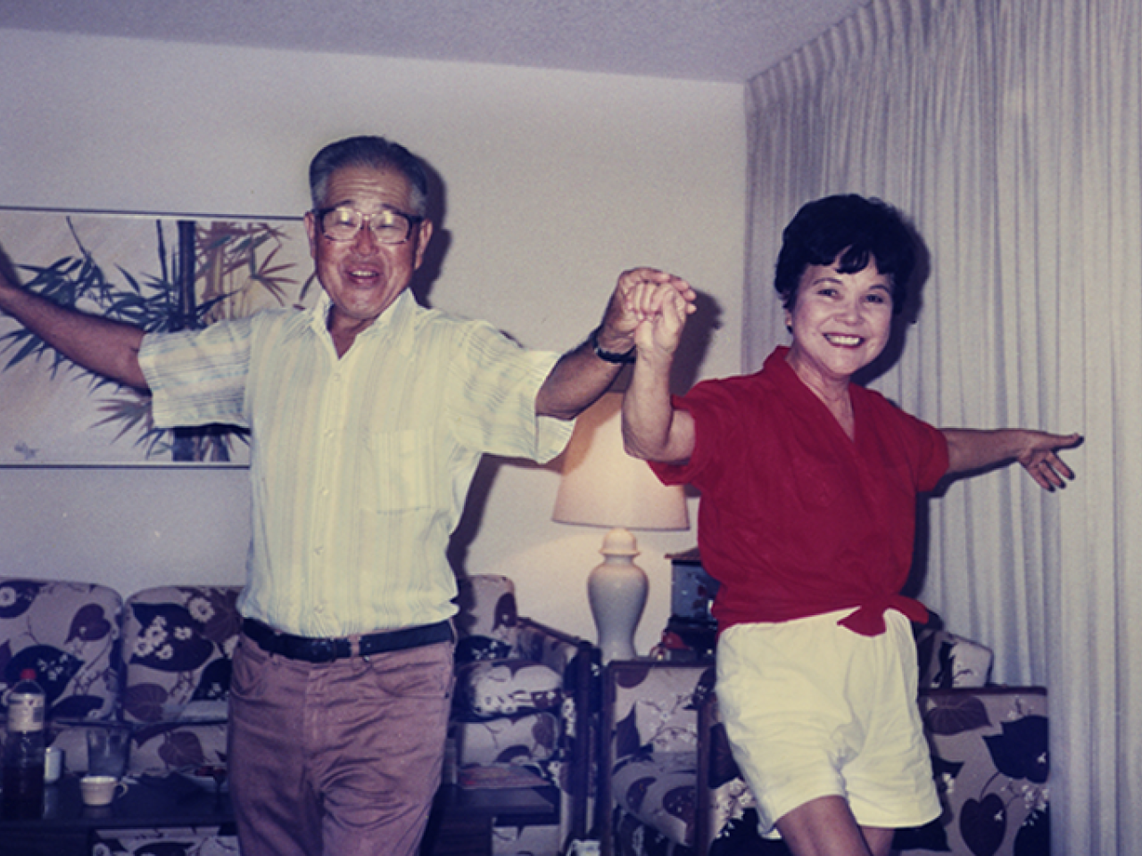 A middle-aged Japanese-American couple hold hands and dance in their living room. Their faces are lit with smiles as they face the camera, arms extended.