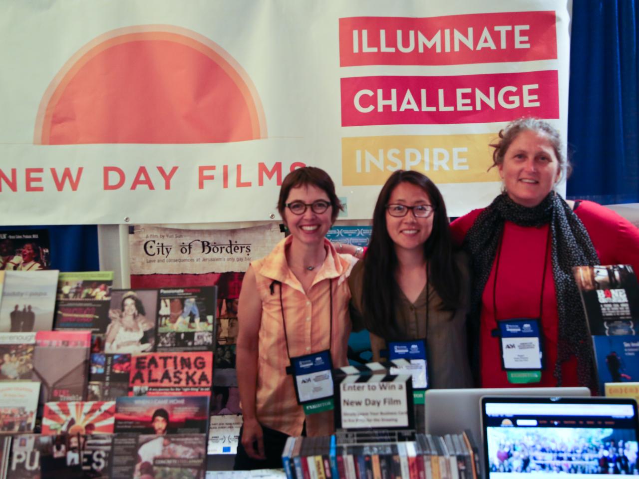 Three New Day filmmakers smile and stand together with conference badges around their necks for the American Sociological Association conference. They’re surrounded by New Day Films including posters, DVDs, a large New Day Films banner, and a laptop with the New Day Films website on it.