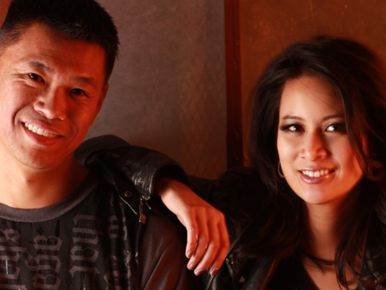 New Day husband and wife filmmaking team Baldwin Chiu and Larissa Lam look directly in the camera smiling softly. Larissa is wearing a leather jacket and has her arm on Baldwin’s shoulder. He is wearing a black shirt with writing on it. They are bathed in red lighting.