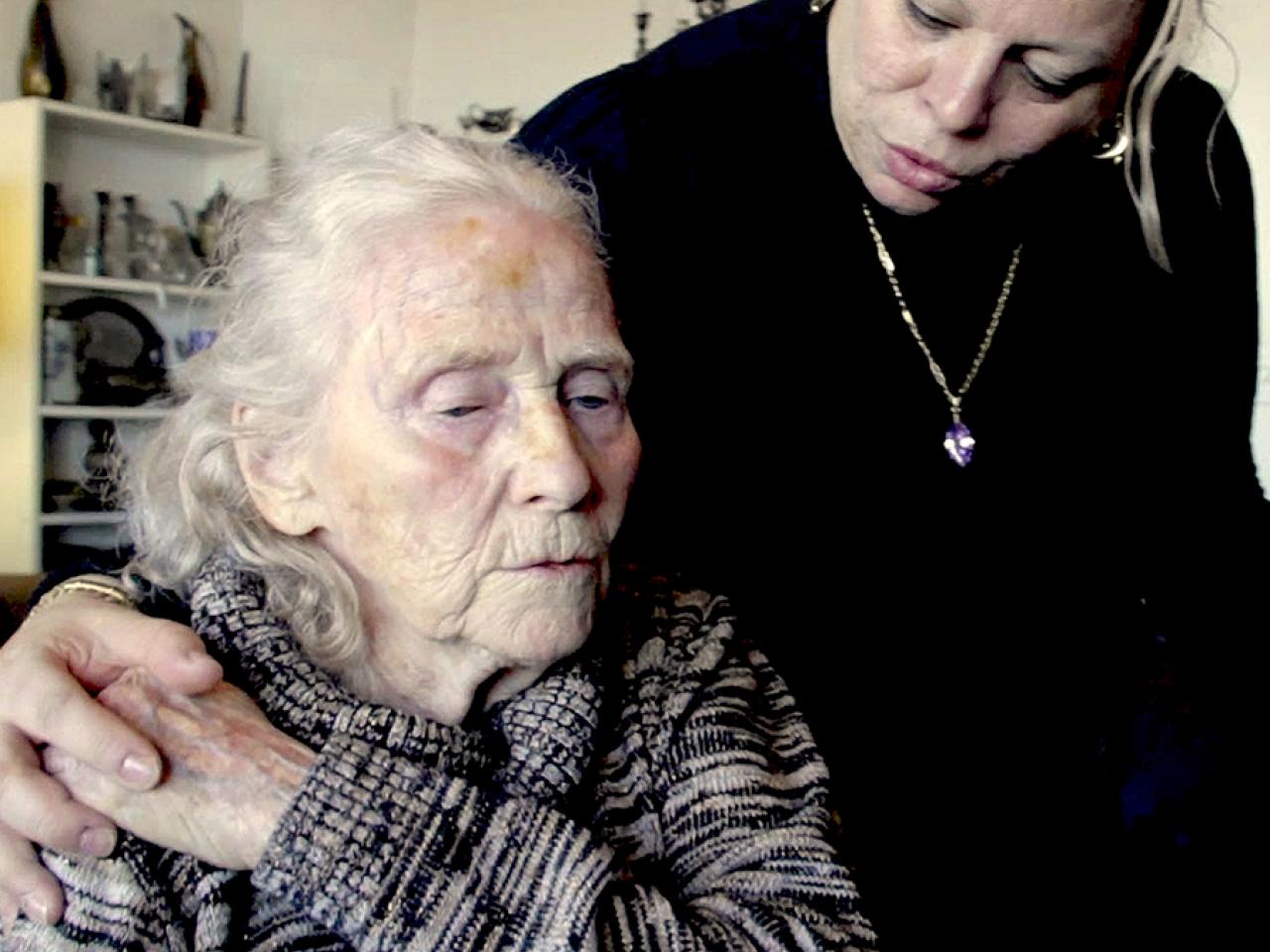 A care worker looks down at an elder woman. They hold hands, and the elder woman's eyes are downcast.