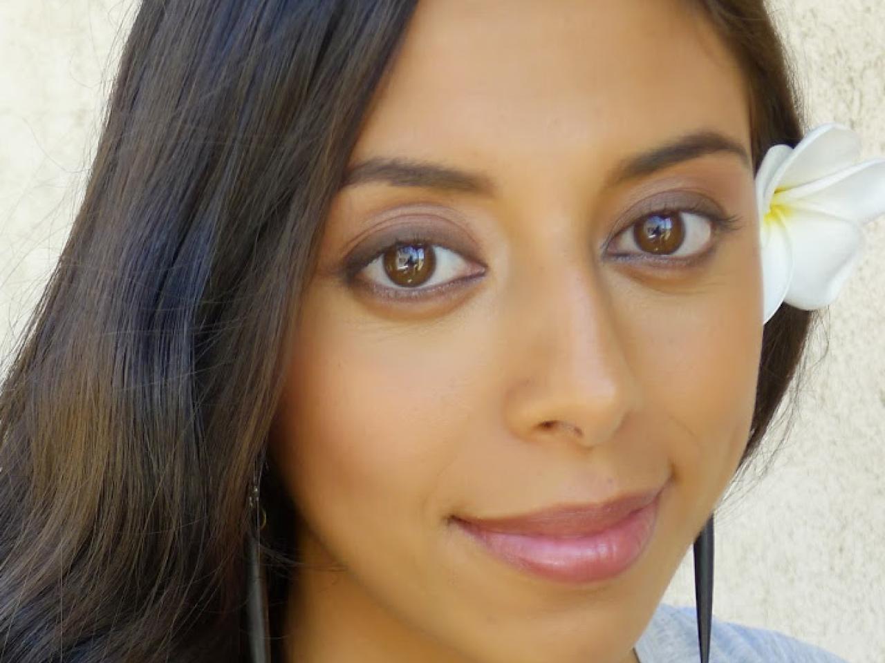 A headshot of New Day filmmaker Chriten Hepuakoa Marquez. A woman with long brown hair and brown eyes wears a white flower tucked behind her ear. She is standing in front of a white background giving a soft, close-mouthed smile to the camera.