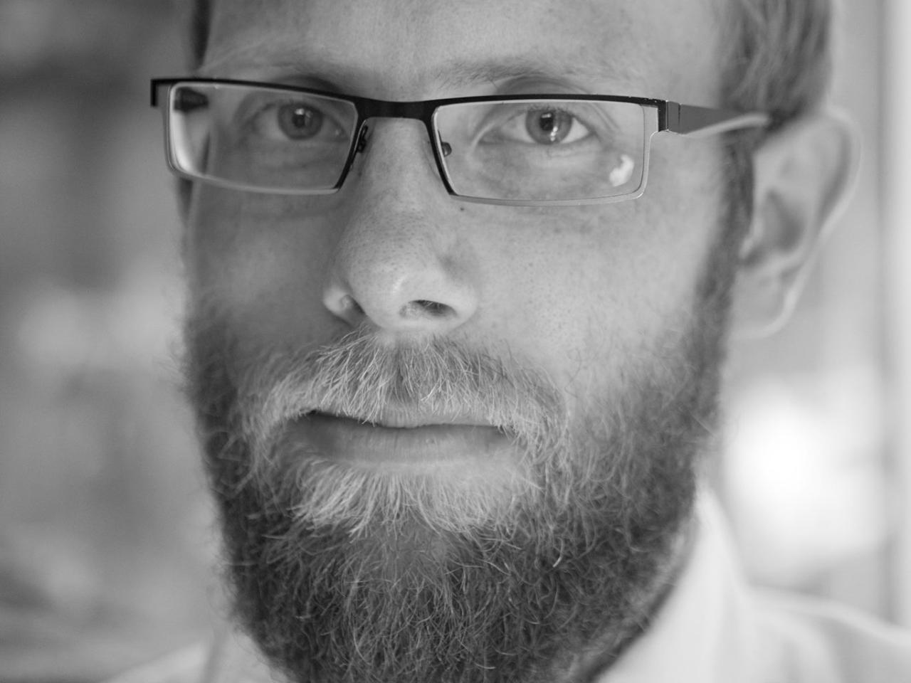 A black and white headshot of New Day filmmaker Christian Jensen. He wears square glasses and a collared shirt and looks into the camera with a serious expression.