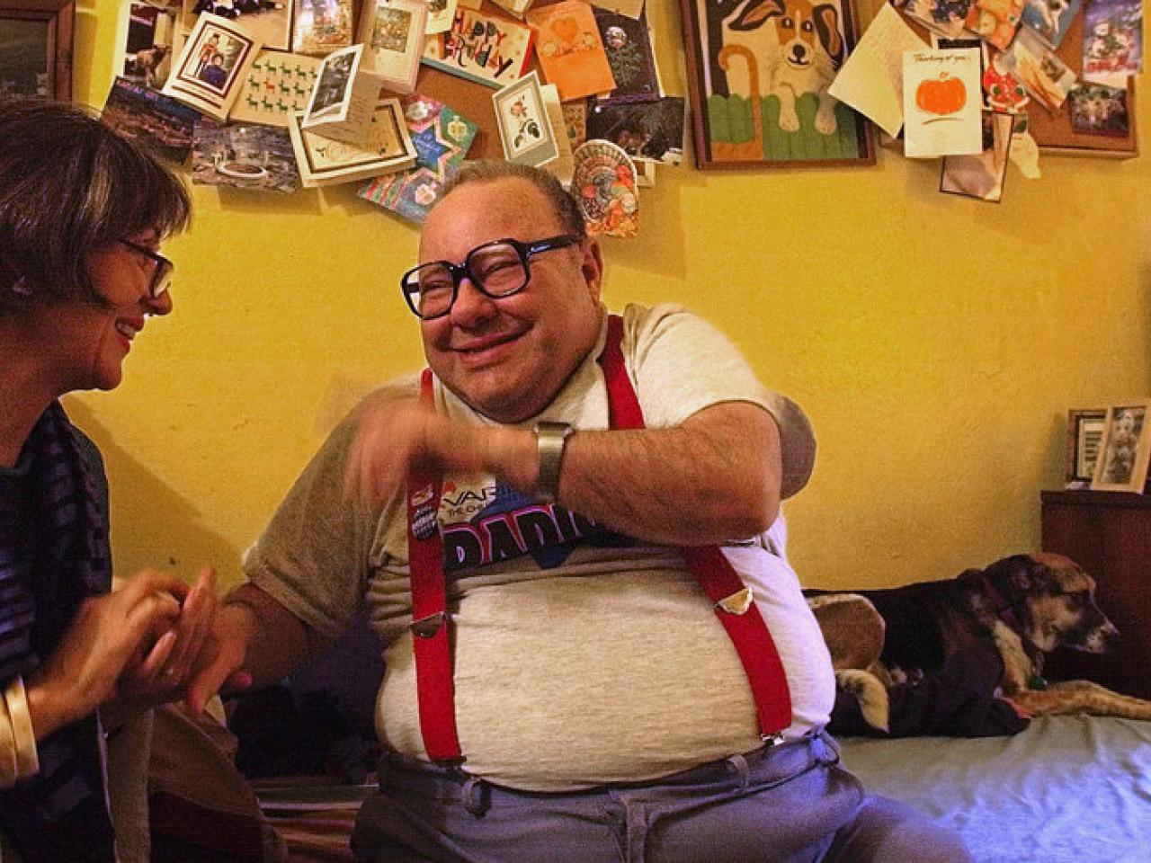 Larry Selman, a community activist and fundraiser with an intellectual disability, and filmmaker Alice Elliot sit, holding hands, talking and laughing in a bedroom. Behind them is a dog, a poster board filled with cards, a dresser with an old TV on top and framed photos.