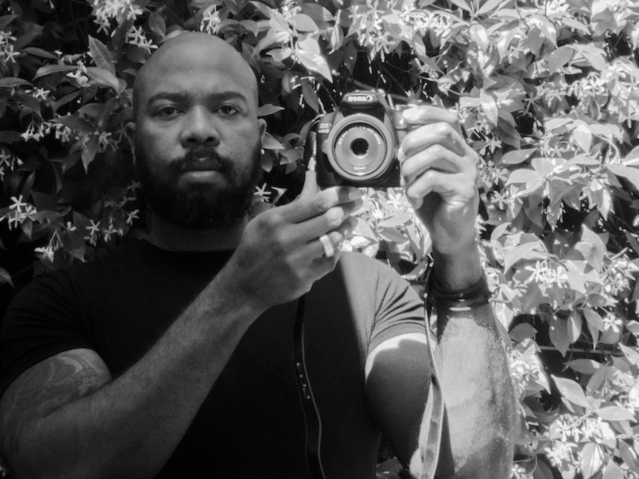 In this black and white photo, African-American filmmaker Daryl P Jones stands in front of flowering jasmine, looking directly at the camera. He holds up a digital video camera near his shoulder. He’s bald and has a thick beard.