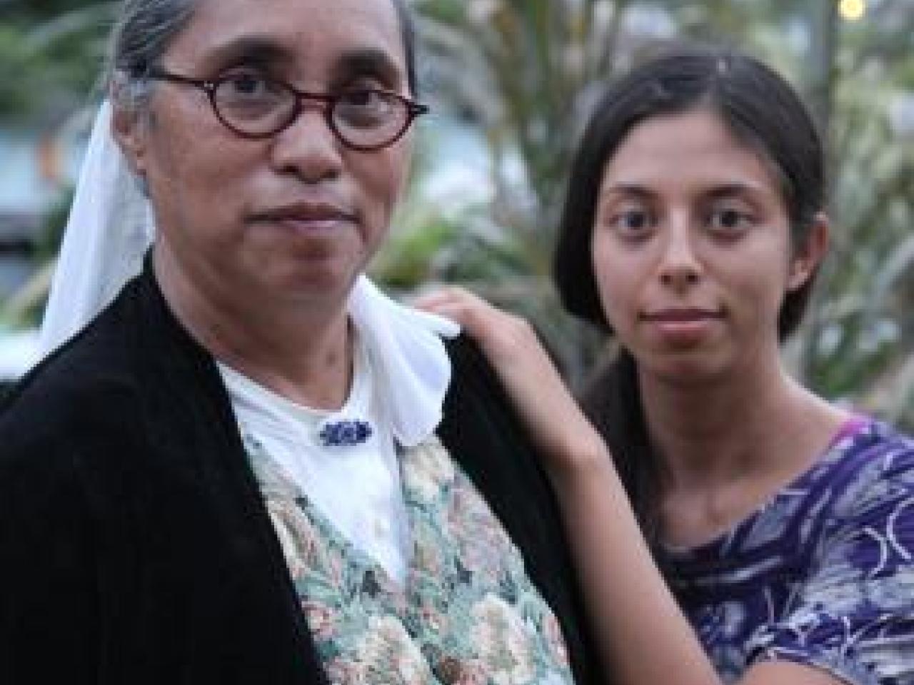 A still from the film E Haku Inoa To Weave a Name. New Day Filmmaker Christen Marquez stands behind her mother and rests her hands on her mother's shoulder. Both women are looking straight into the camera with similar faint smiles on their faces. Lush greenery fills the background.