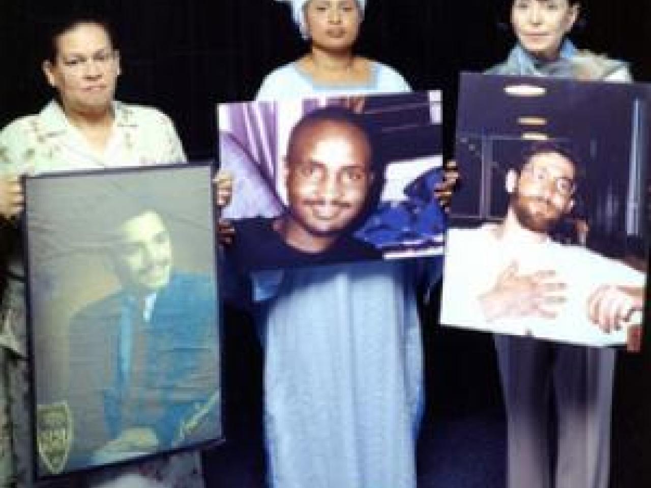 Three women stand shoulder to shoulder and look solemnly at the camera. The each hold a large photo of their late sons.