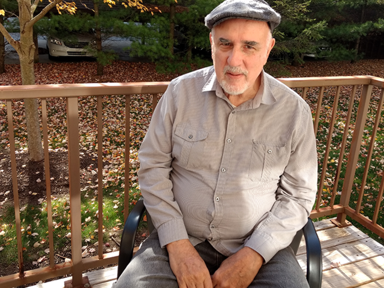 New Day Member George Bogdanich, a white man with a gray goatee, sits on a black chair and looks up at the camera. Behind him is a wooden porch railing and green grass dotted with fallen autumn leaves.