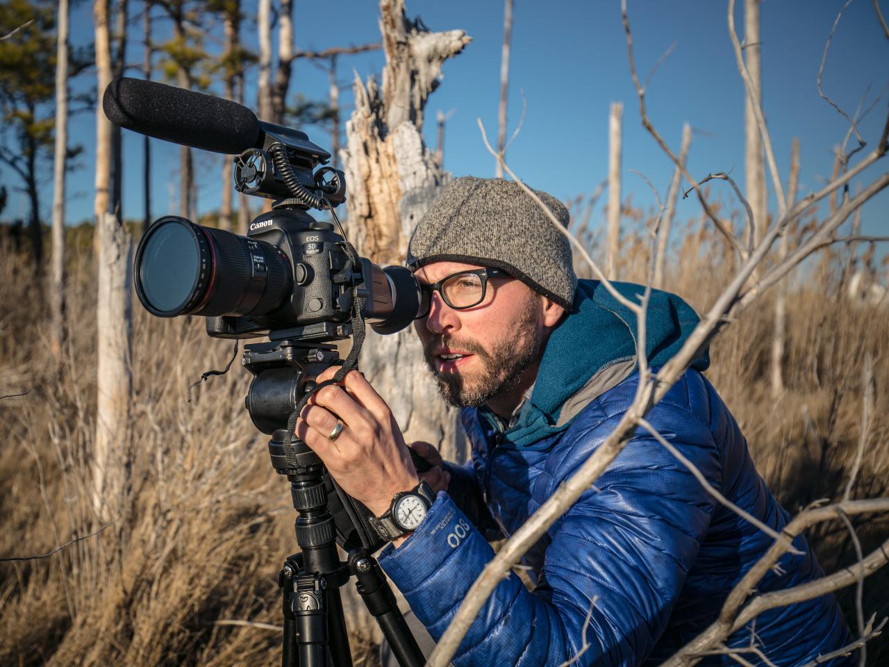 New Day Member Michael O. Synder crouches in a field and looks through a camera that’s mounted on a tripod. The ground is brown, the trees are bare, and Michael wears a blue winter jacket.