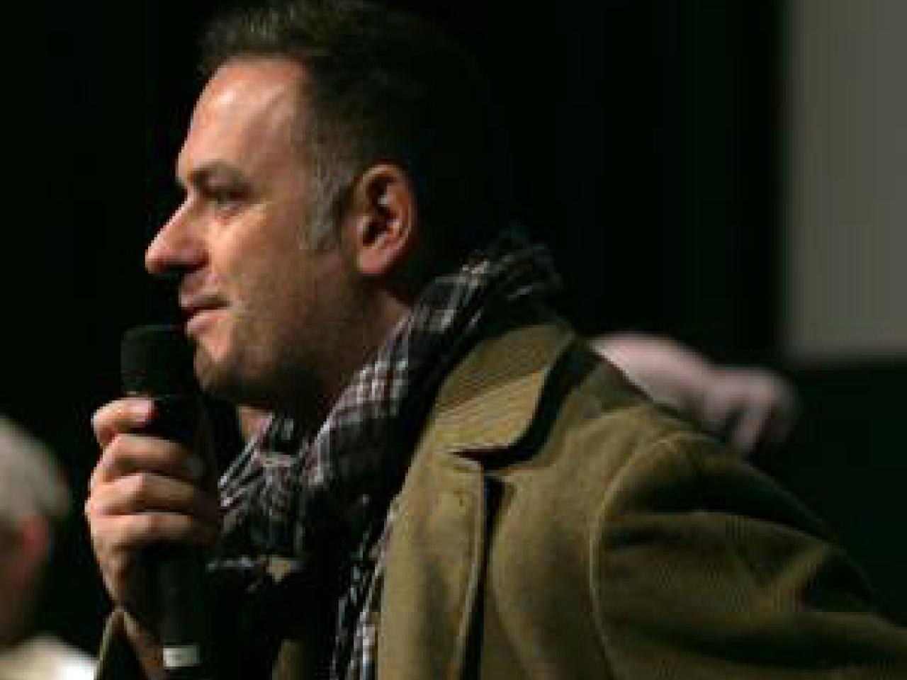 A side shot of New Day filmmaker Jean-Michel Dissard. He wears an olive green jacket and a plaid scarf. He holds a microphone to his mouth with an amused expression on his face.