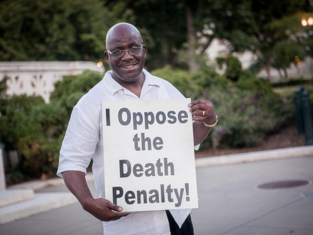 Jerry Givens smiles, holding a sign with "I oppose the death penalty!"