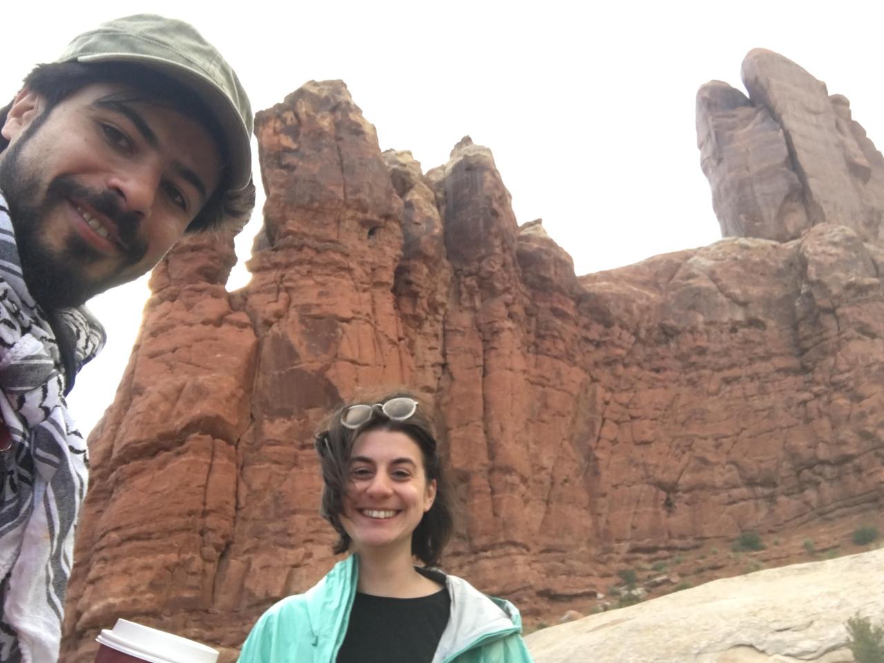 New Day Film directors Jillian Karole and Ahmed Mansour smile at the camera on a windy day. They stand outside with a rocky, sandy cliff towering behind them.