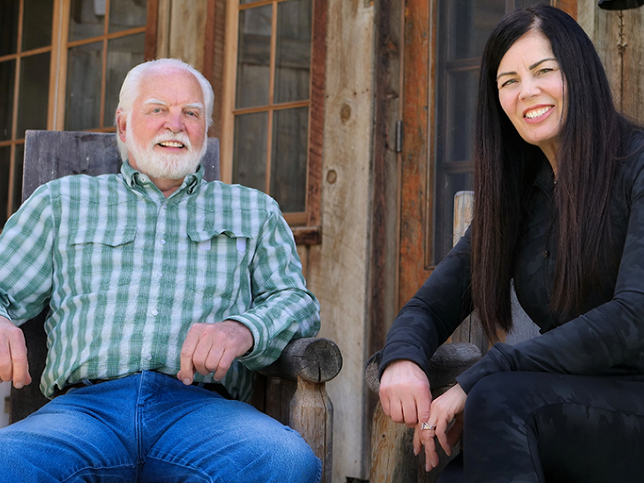 New Day Filmmakers Joe Phelps and Rosemary Smith sit side by side on wooden chairs, smiling at the camera. Joe wears a green and white plaid shirt and Rosemary is dressed in black.