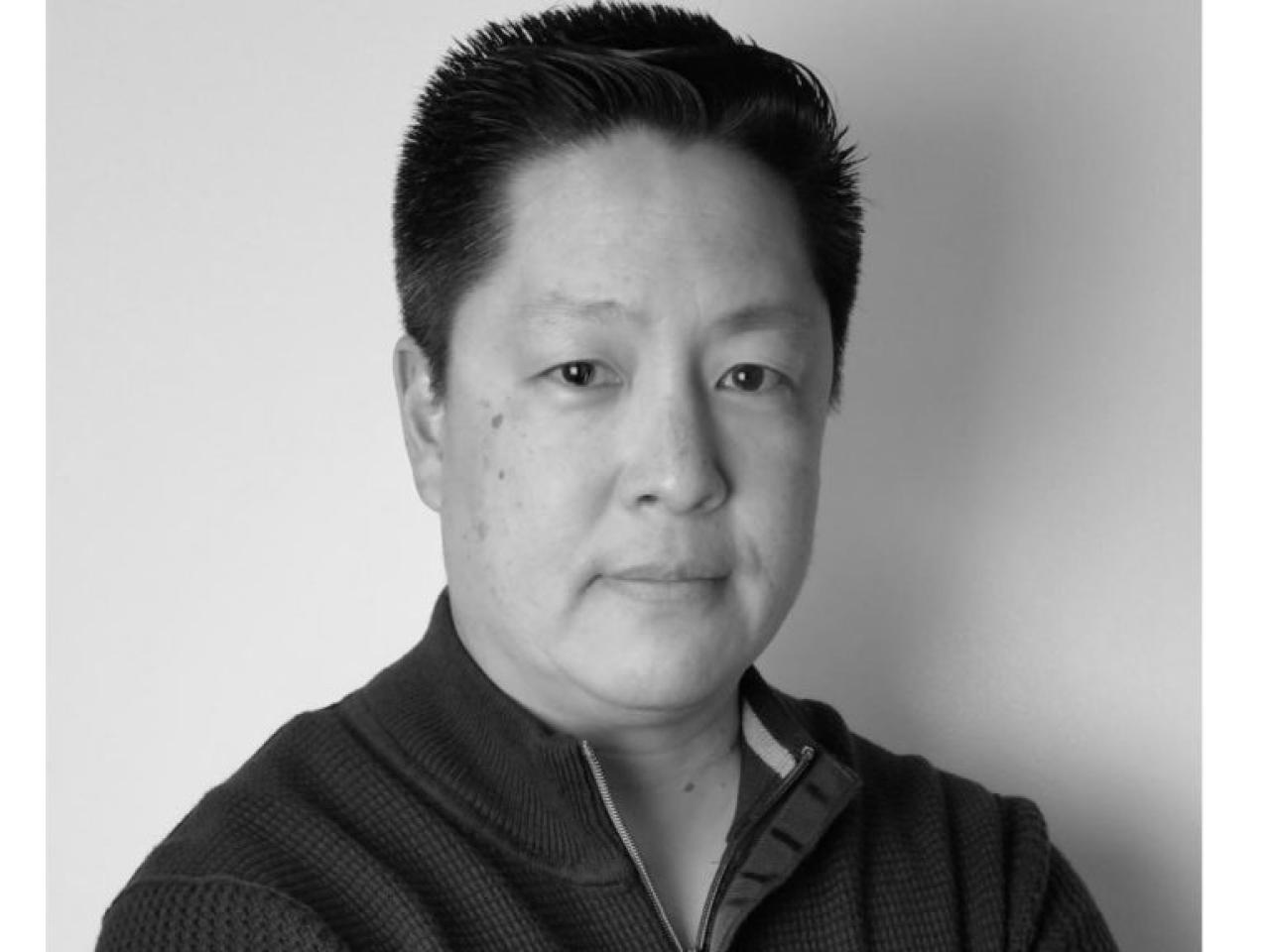 New Day Director Jon Osaki, a Japanese American, stares directly into the camera in this black and white headshot.