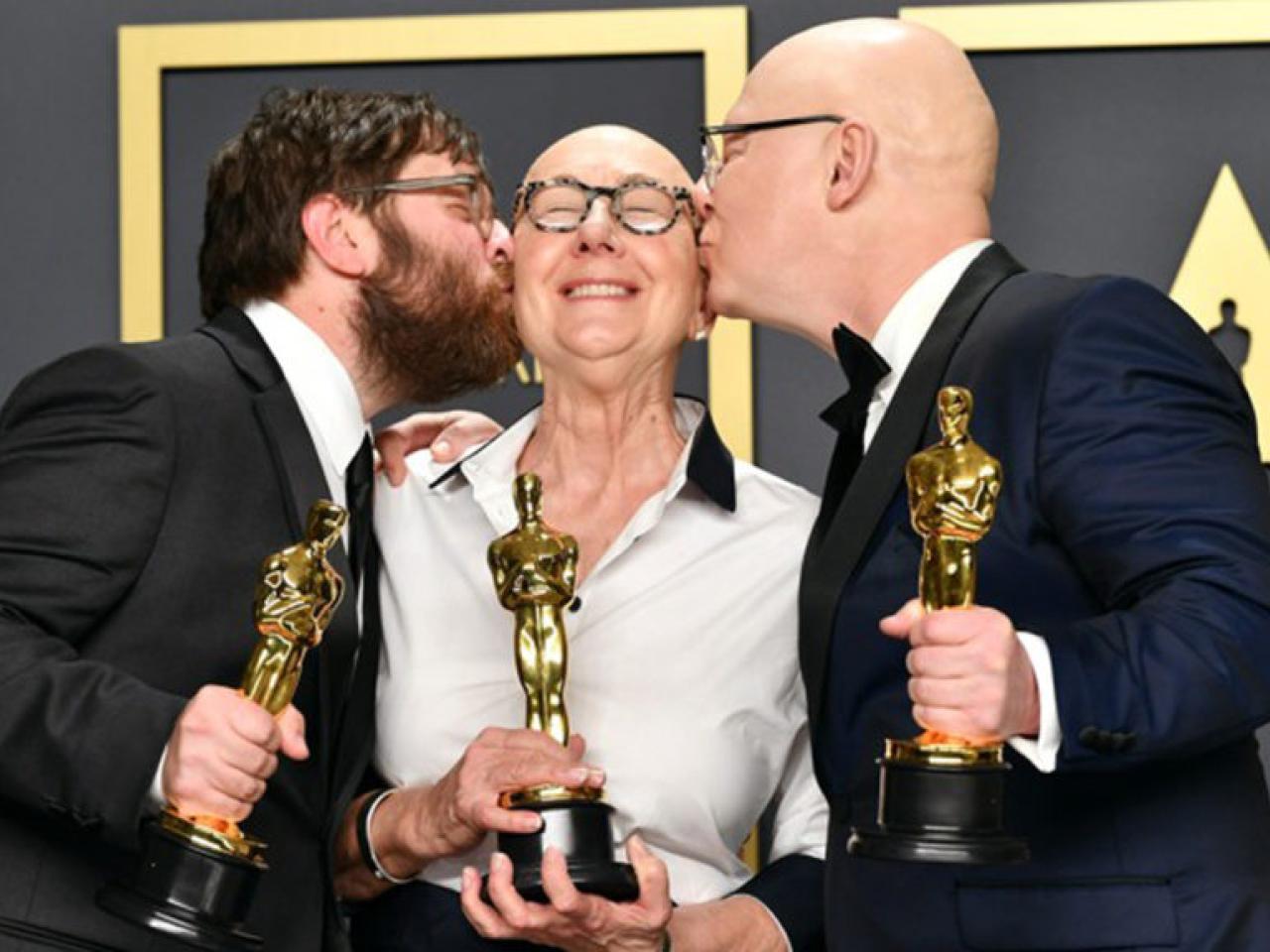 Julia Reichart, with a bald head and round glasses, holds an Oscar. She smiles with her eyes closed, and is kissed the men on either side of her, each holding an Oscar statue.
