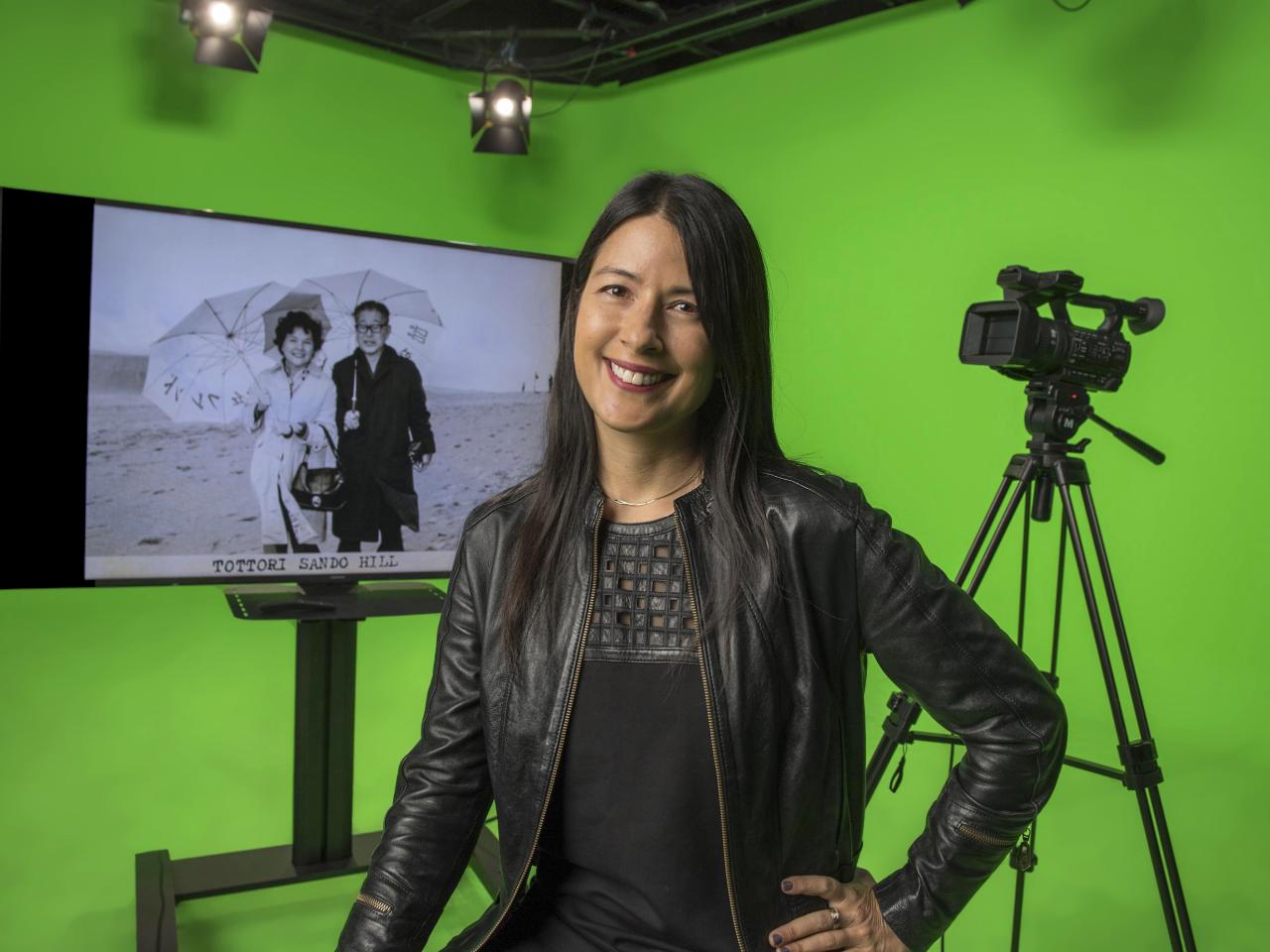 Kimi Takesue smiles for the camera in a studio with green-screen walls. Behind her, a video camera on a tripod and a monitor showing a black and white image of her grandparents on the beach with parasols.
