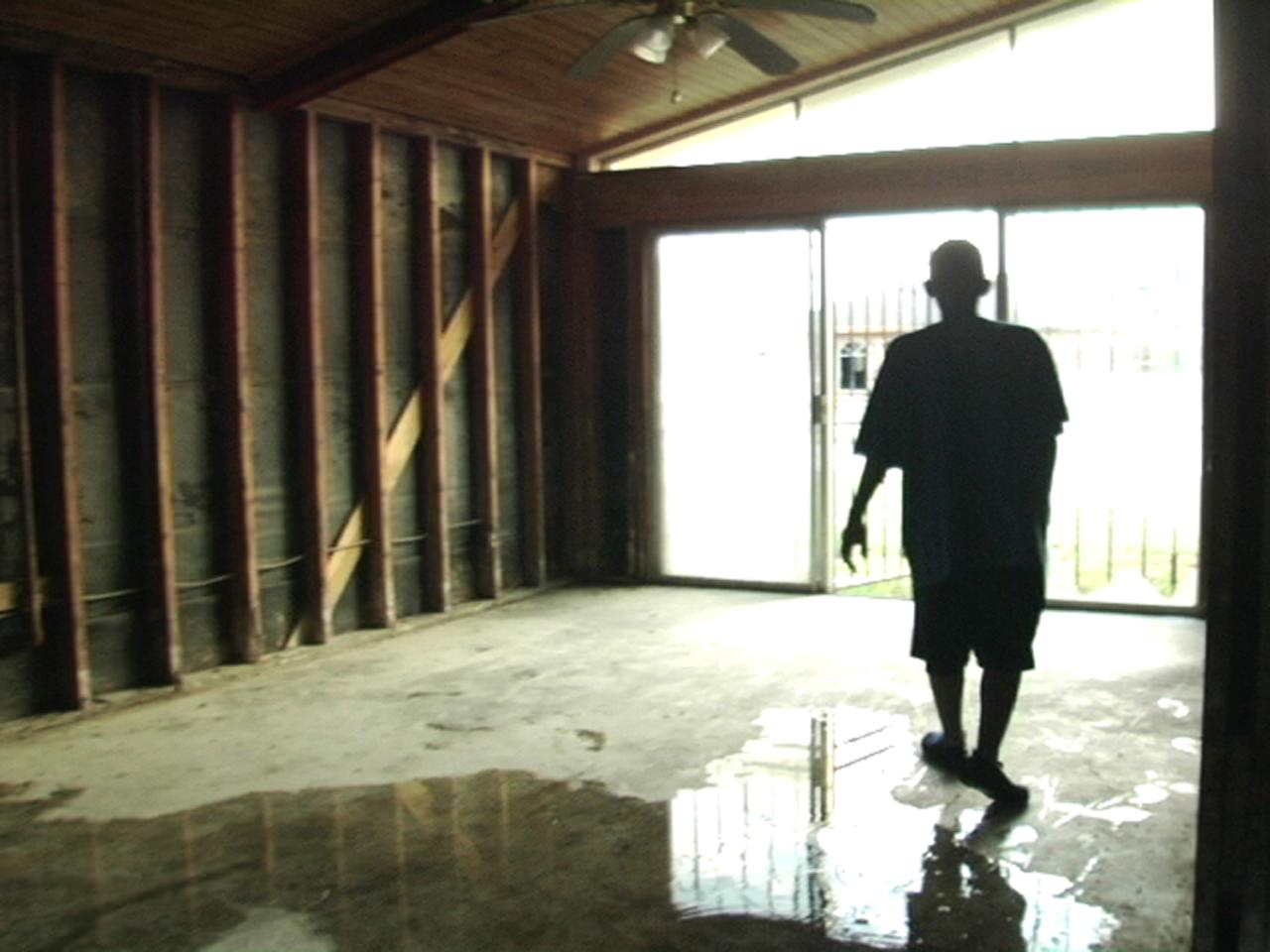 A man stands inside a building where the wall beams are fully exposed and water pools on the cement floor. He is silhouetted against bright windows in the background.