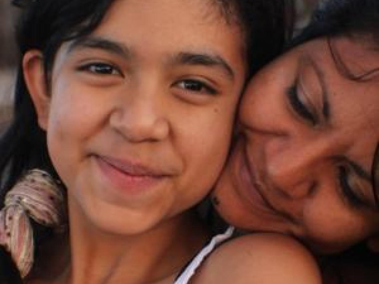 A still from the New Day film Life on the Line. A young Mexican-American girl, Kimberly Torrez smiles sweetly towards the camera. A woman, likely her mother Vanessa Torrez sits behind Kimberly and rests her head on her shoulder. She turns her face into Kimberly’s neck with a loving smile.