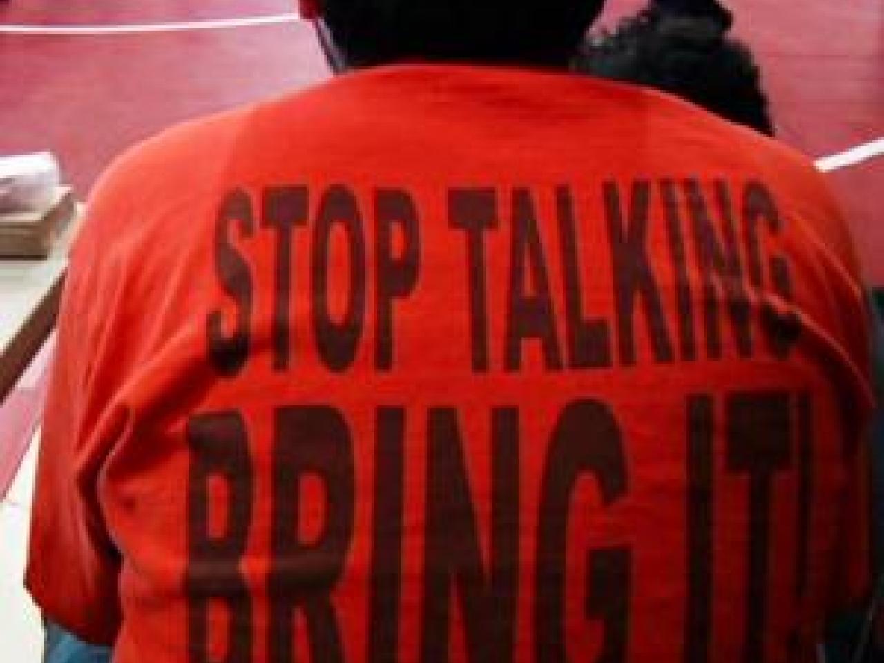 A still from the New Day film Making Noise In Silence. The back of a person sitting on a bleacher. They are wearing a bright orange t-shirt that reads “Stop Talking, Bring It!” in large black letters. The ground is a red gymnasium floor with a wrestling ring.