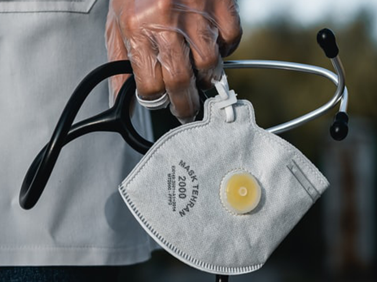A latex-gloved hand holding a disposable respirator and a stethoscope. The hand is next to the torso of the person's white lab coat.