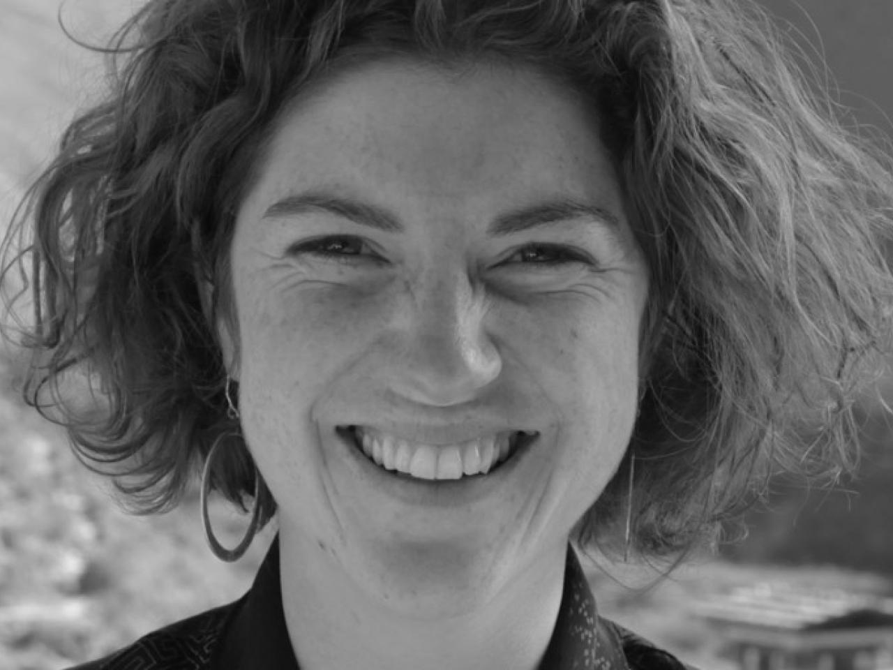 In this black and white photo, New Day filmmaker Meghan Shea looks directly at the camera and smiles widely. Her medium-length hair is above her shoulder and she wears hoop earrings.