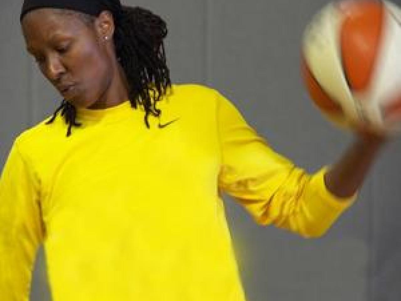 Chamique Holdsclaw, an African-American woman with her locs pulled back looks intently toward the ground, lips pursed. She wears a bright yellow Nike long-sleeved shirt and holds a basketball aloft in one hand.
