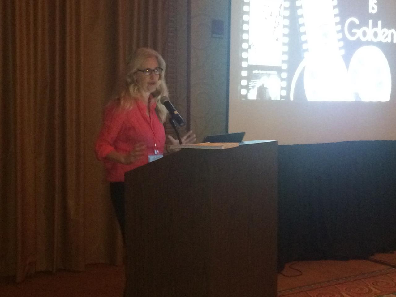 Laurel Chiten wears a bright pink blazer and stands in front of a podium on the stage at the New Day House in the National Media Market. There is a bright screen on her left with an image of film strips on it.