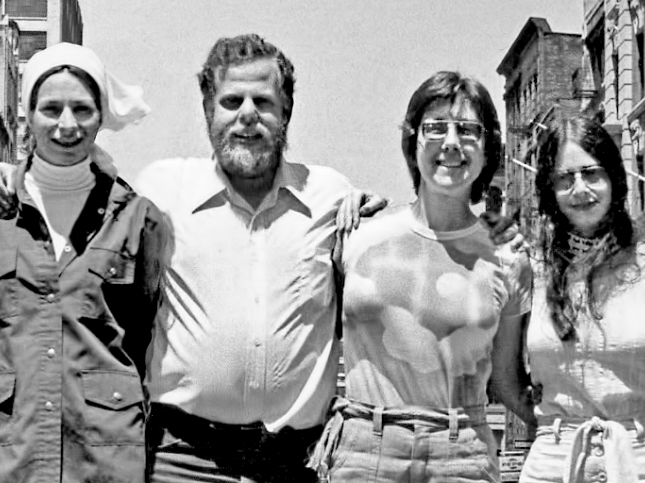 In this black and white shot from the 1970s, New Day Co-founders link arms and smile at the camera. From left to right:  Liane Brandon, Jim Klein, Julia Reichert, and Amalie R. Rothschild.