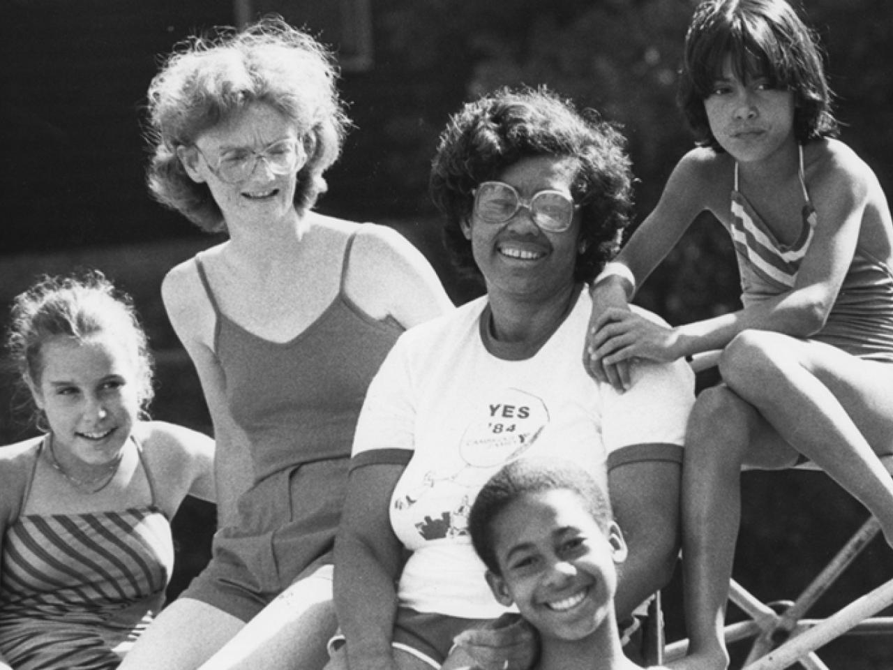 In this black and white photo from the 1980s, five teens and children of various races sit on playground equipment smiling on a bright summer’s day.