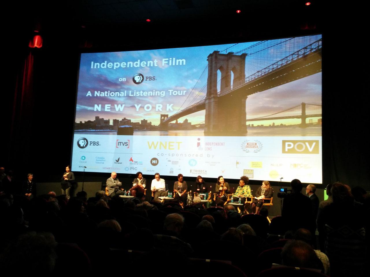 An auditorium with a large screen on the stage. On the screen is an image of Brooklyn Bridge and the words “Independent Film on PBS A National Listening Tour New York” in white text. On stage a panel of people of various races sit in a row, some leaning over in discussion with one another.