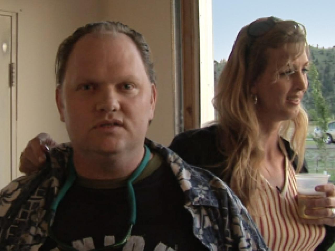 A still from the New Day film Prodigal Sons. A young man faces the camera with a slightly surprised expression as if he wasn’t expecting a picture to be taken. His sister, New Day filmmaker Kimberly Reed rests her hand on his shoulder. She is standing behind him and facing to the side with a drink in her hand. Behind her is a window with trees outside.