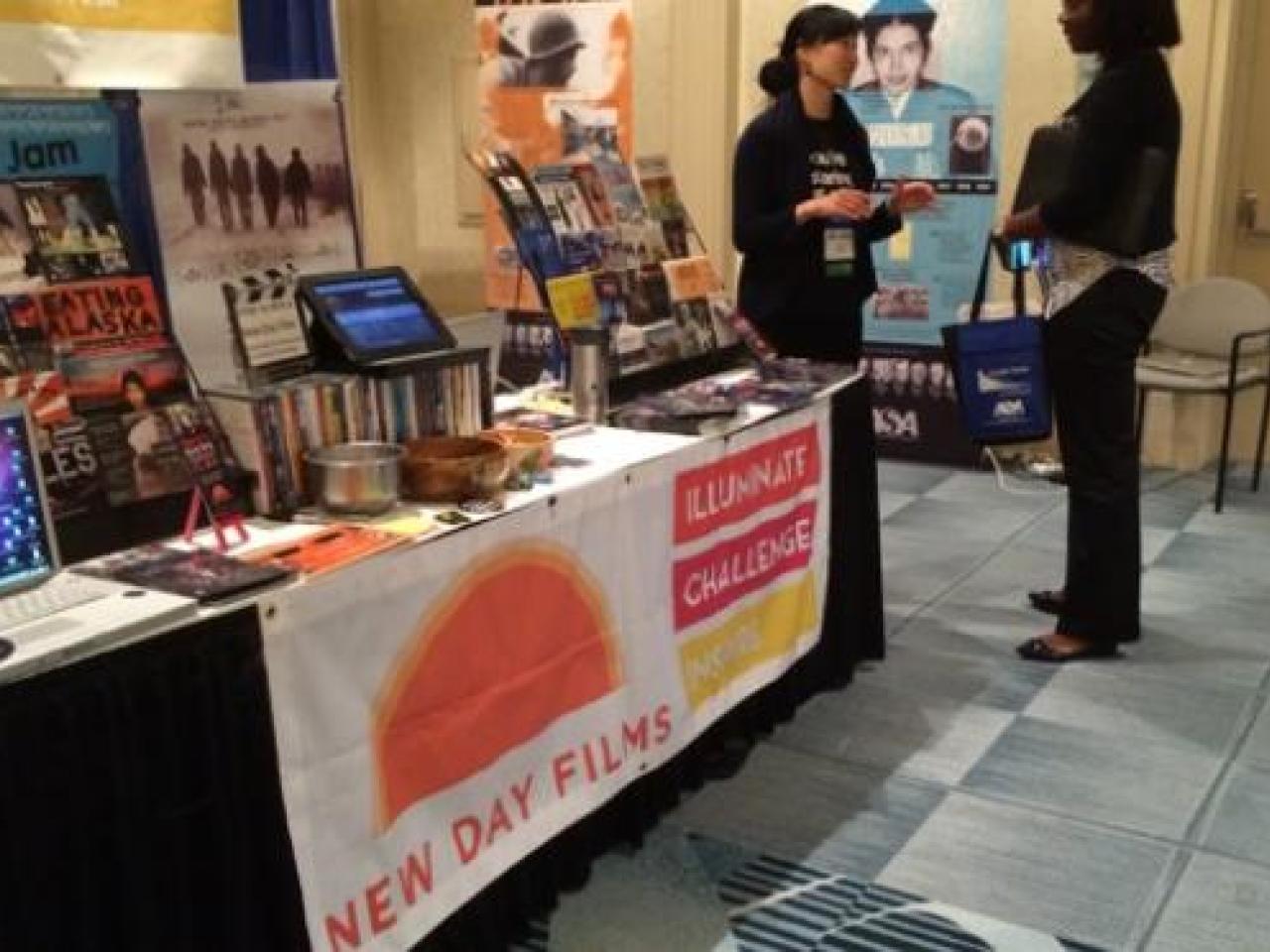 New Day filmmaker Debbie Lum converses with an attendee at the New Day exhibition booth filled New Day films promotional materials and a laptop to screen trailers.