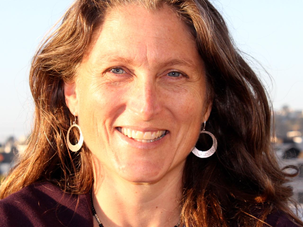 A headshot of New Day filmmaker Reagan Brashear. She smiles and looks into the camera with the sun shining across her face. She wears long earrings with silver circles at the bottom. In the background is a light-blue sky.