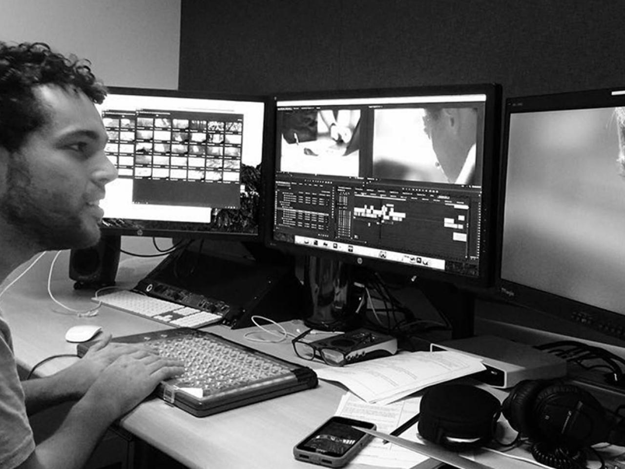 New Day Filmmaker Reid Davenport is sitting at a desk with his hands on a keyboard in a video editing room. He faces three video monitors and has a small smile on his face.