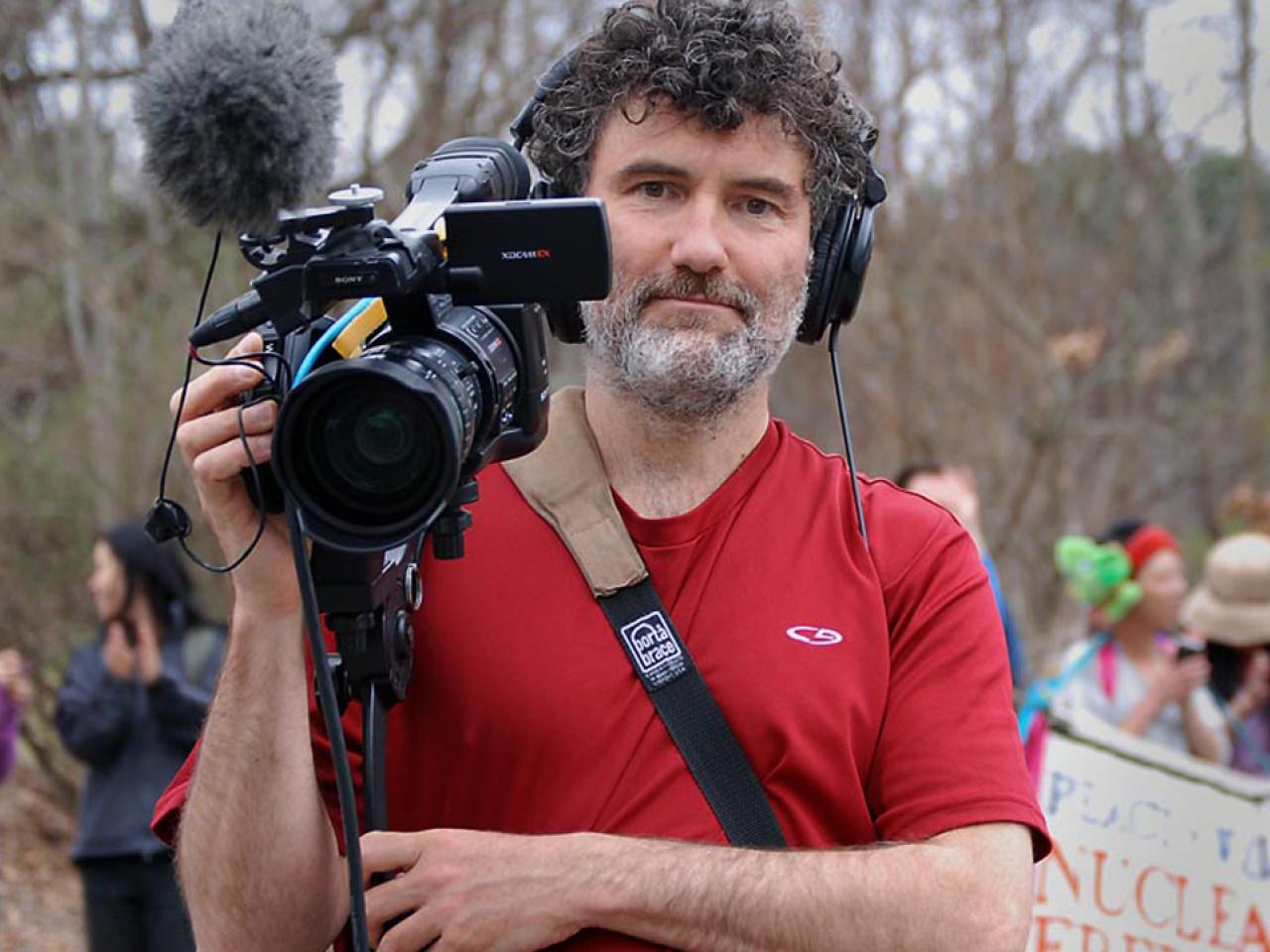 Robbie Leppzer on location filming at a protest holds his video camera and wear headphones.