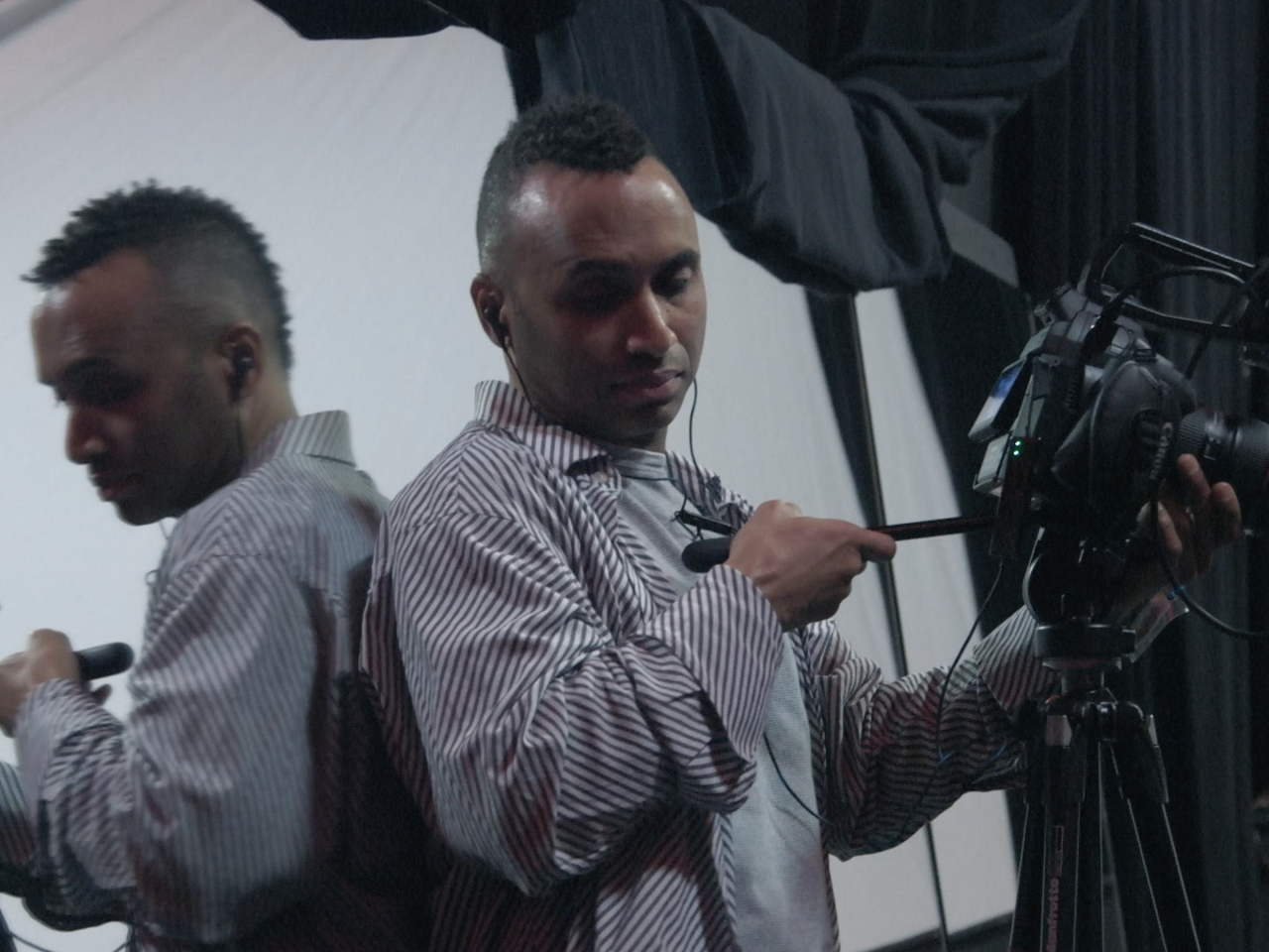 Rodney Evans, a Black man, focuses a video camera on a tripod while looking down into the camera's monitor. He leans against a mirrored wall, his face and the camera reflecting back.