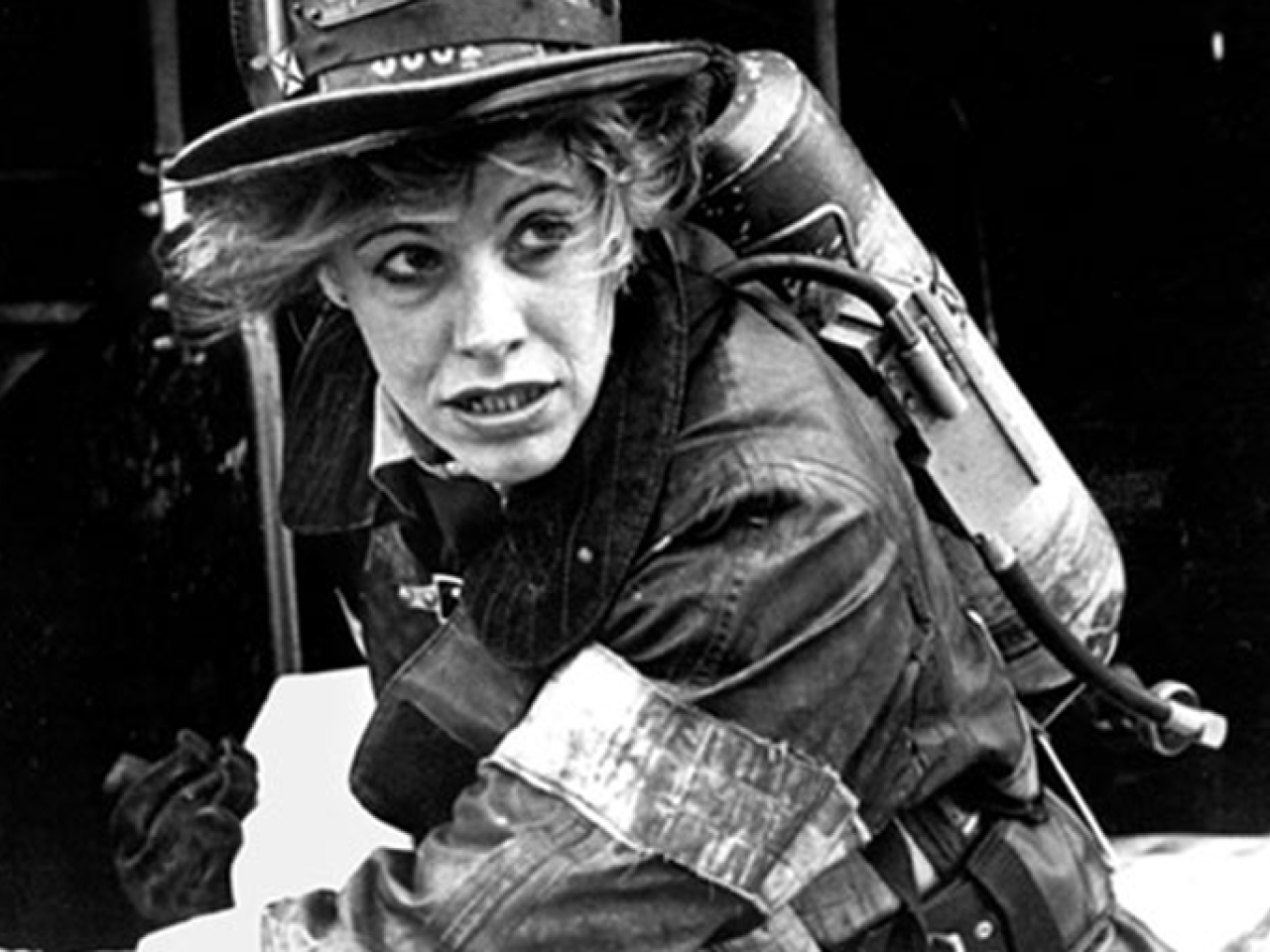 In this black and white still from the film, a female firefighter is fully geared up in a heavy coat, gloves and helmet as she races at the scene of a fire. She wears an oxygen tank on her back and looks out towards the distance, with determination on her face.