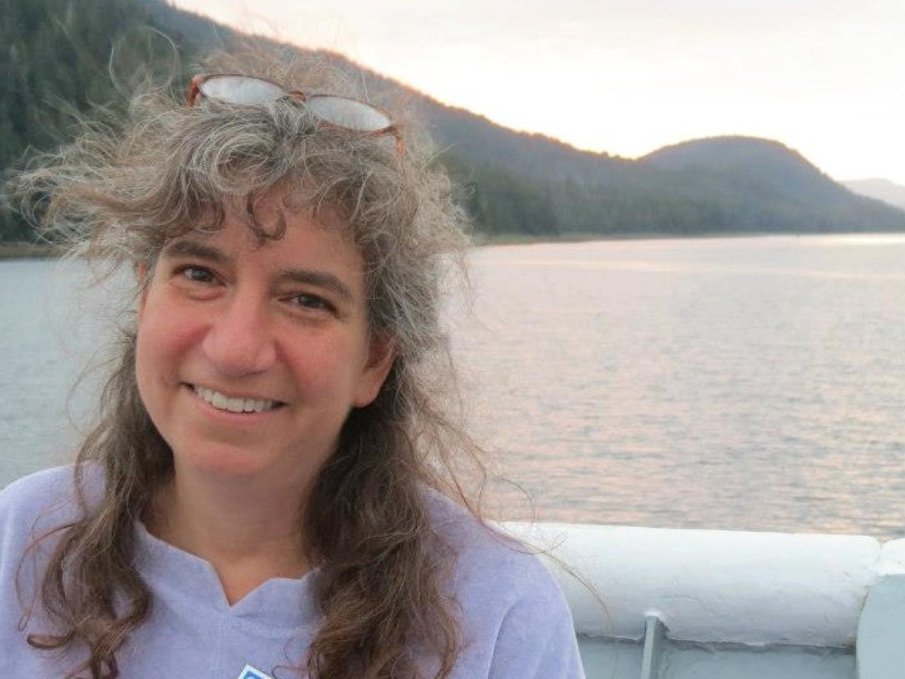 A headshot of New Day filmmaker Ellen Brodsky. Ellen smiles and stands on a boat with the ocean and mountains in the background. Her glasses are on top of her hair which is blowing in the breeze.