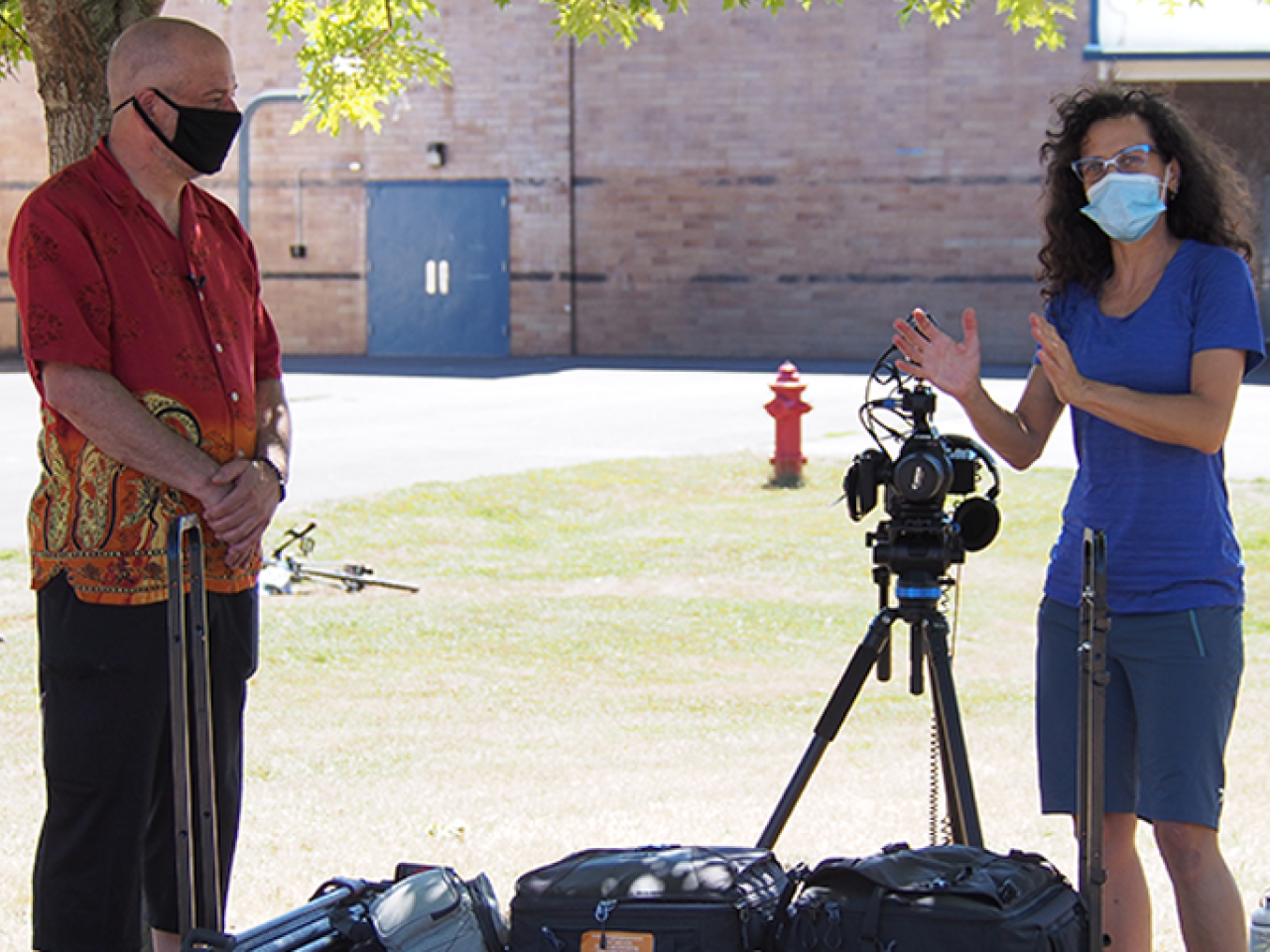 New Day Member Cheryl Green stands in a small patch of shade at an outdoor film shoot with Karl, the star of “TBI & My Longest Ride.” They are white, and both wear masks, standing at opposite ends of a cart full of video gear. Cheryl, by her camera and tripod, looks toward the distance and gestures as Karl watches her discuss the next shot.