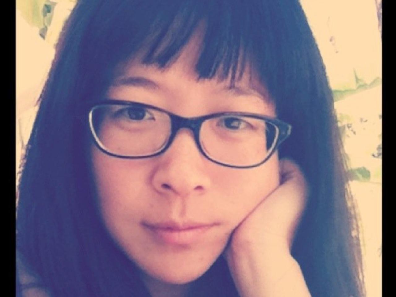A headshot of New Day filmmaker Mina T. Son, a Korean-American woman with bangs and glasses. She rests her palm on her cheek in a thoughtful pose and looks into the camera. Around the edges of the photo is a black frame with letters and numbers in orange on top as if the photo were a digital photo.